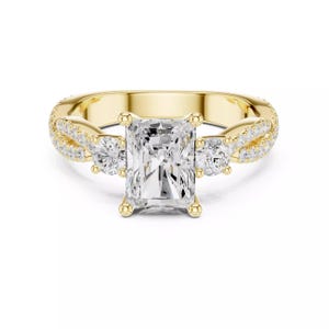 Op de afbeelding: Een gouden ring met een rechthoekige diamant in het midden. Twee kleinere, ronde diamanten flankeren de hoofdsteen. De band heeft een gedraaid ontwerp met kleinere, ingebedde diamanten.