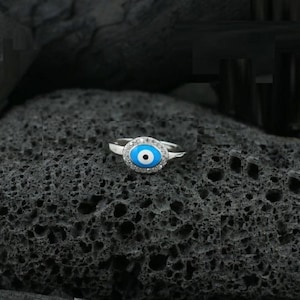 May include: A silver ring featuring a blue and white evil eye design, surrounded by small, clear gemstones. The ring is set against a dark, textured background, highlighting the ring's details.