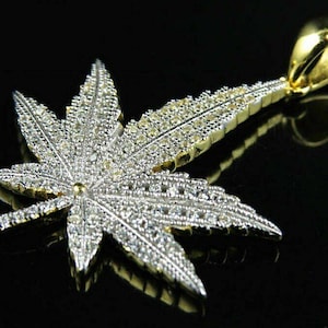 Puede incluir: Un colgante de hoja de marihuana de oro y plata con piedras de circonita cúbica transparentes.