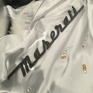 May include: A white satin fabric with the word "Maserati" spelled out in black rhinestones. The fabric is draped over a black surface and there are several small, clear rhinestones scattered around the fabric.