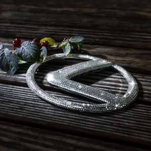 May include: A silver car emblem with a diamond-like finish, shaped like a stylised letter K, on a dark wooden surface.