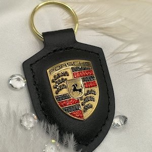 May include: Black leather keychain with a gold Porsche logo featuring red, black, and silver rhinestones. The logo says "PORSCHE" and "STUTTGART".