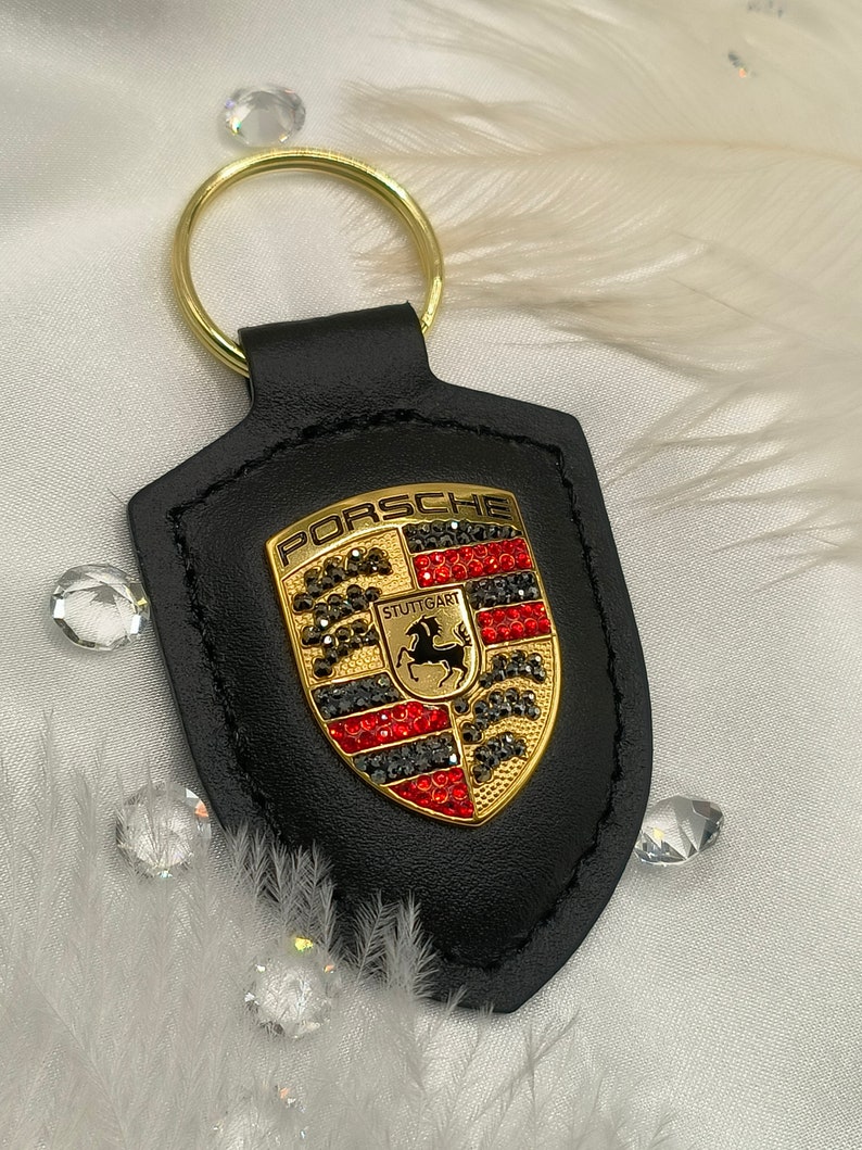 May include: Black leather keychain with a gold Porsche logo featuring red, black, and silver rhinestones. The logo says "PORSCHE" and "STUTTGART".