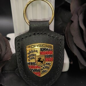 May include: Black leather keychain with a gold Porsche logo featuring red and black rhinestones. The logo says "Porsche" and "Stuttgart".