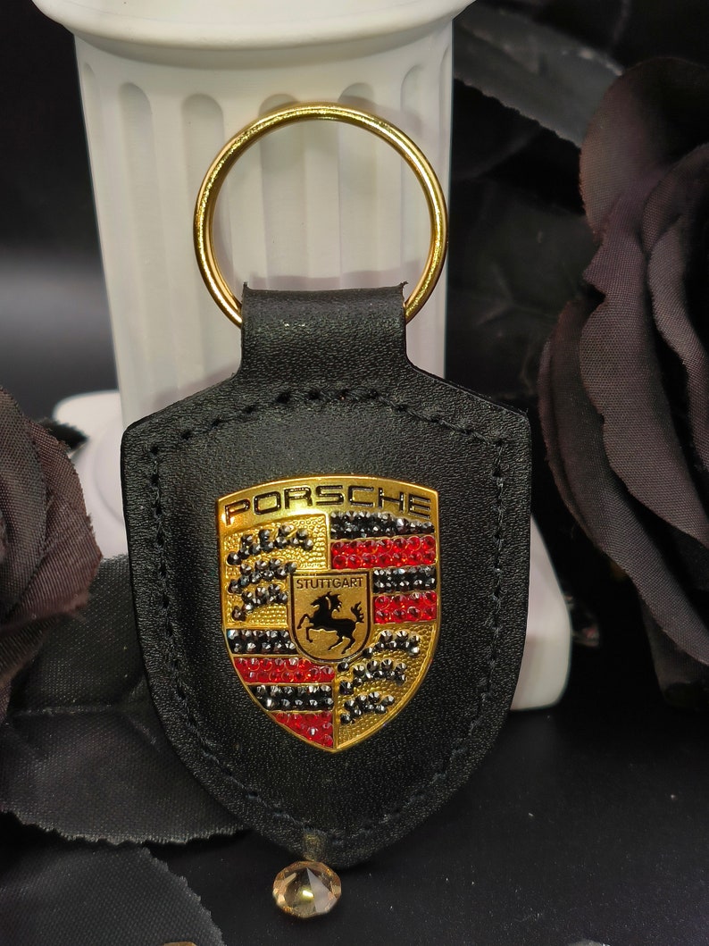 May include: Black leather keychain with a gold Porsche logo featuring red and black rhinestones. The logo says "Porsche" and "Stuttgart".