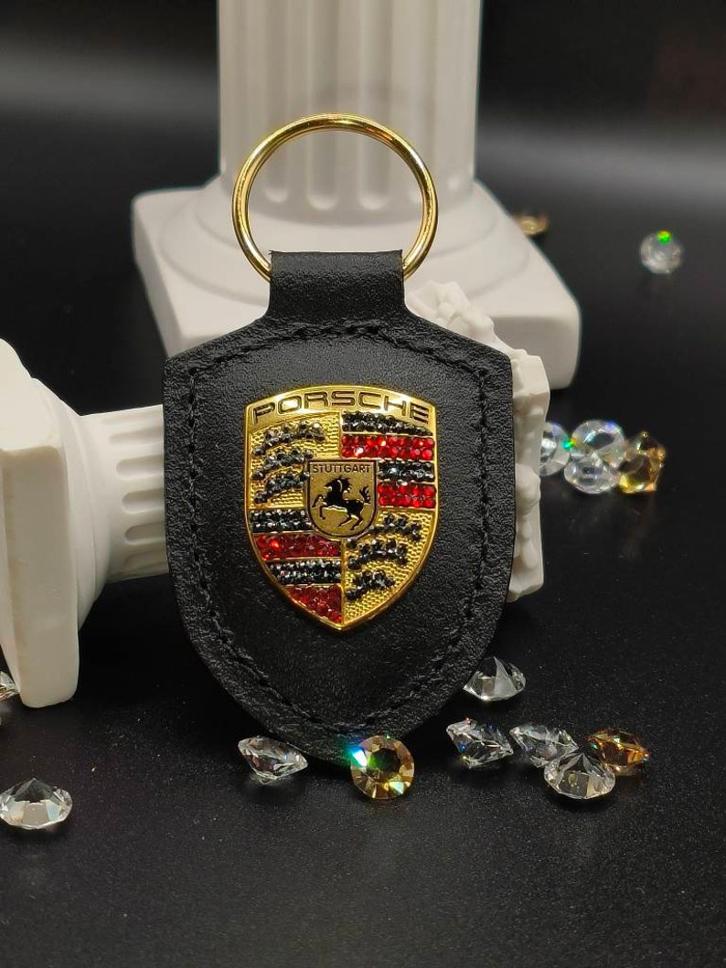 May include: Black leather keychain with a gold Porsche logo featuring red, black, and white rhinestones. The logo says "Porsche" and "Stuttgart".