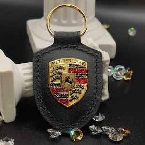 May include: Black leather keychain with a gold Porsche logo featuring red and black rhinestones. The logo says "PORSCHE" and "STUTTGART".