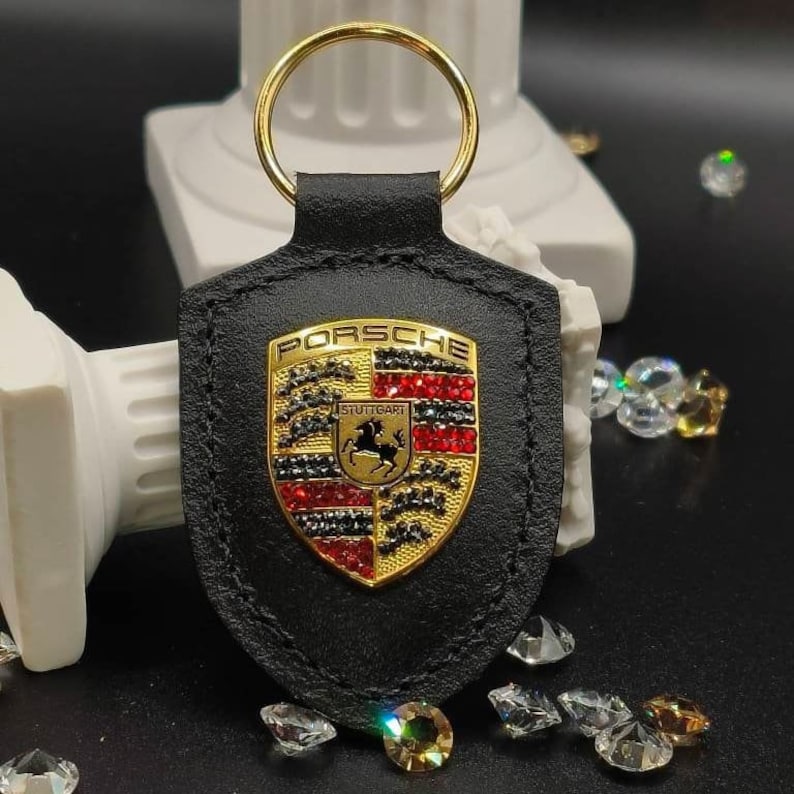 May include: Black leather keychain with a gold Porsche logo featuring red and black rhinestones. The logo says "PORSCHE" and "STUTTGART".