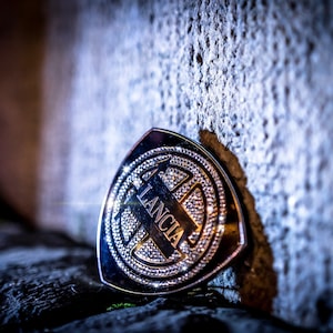 May include: A silver and diamond encrusted car emblem with the word "LANCIA" in a stylised font.