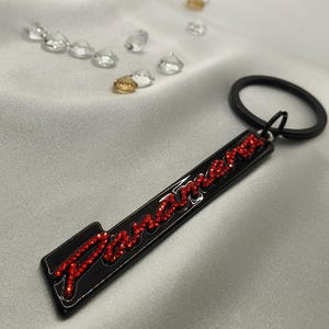 May include: Black rectangular keychain with the word "Panamera" in red rhinestones. The keychain has a black ring and is set against a white satin background with scattered clear and gold crystals.