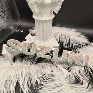 May include: White, sparkly, rhinestone letters spelling out the word "SUZUKI" on a black background with white feathers.