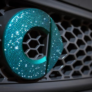 May include: A close-up of a teal blue car emblem with a diamond-like texture. The emblem is on a grey honeycomb-patterned grille.