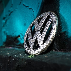 May include: A silver Volkswagen logo made of rhinestones, set against a teal background.
