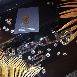 May include: A black box with a gold leaf design and the word "Lexus" spelled out in black crystals. The box has a black card on top with gold lettering that reads "Black Carpet Crystals" and "Jewellery for cars and more with Crystals from Swarovski."