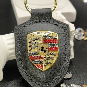 May include: Black leather keychain with a gold Porsche logo featuring red and black rhinestones. The logo says "PORSCHE" and "STUTTGART".
