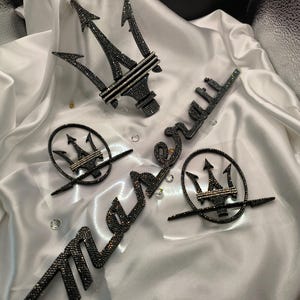以下が含まれることがあります： ラインストーンで飾られた黒と銀の Maserati ロゴが 3 つ。ロゴは三叉の形状で、"Maserati" という言葉はラインストーンで書かれています。