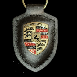 May include: A black leather keychain with a gold Porsche crest featuring a black horse and red, black, and white accents. The crest is surrounded by black rhinestones.