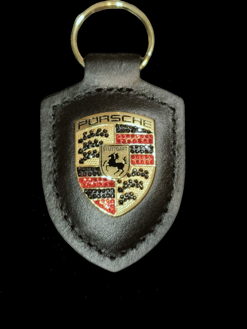 May include: A black leather keychain with a gold Porsche crest featuring a black horse and red, black, and white accents. The crest is surrounded by black rhinestones.