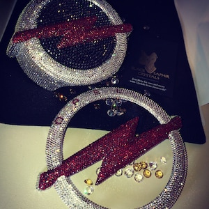 May include: Two silver and red crystal-encrusted car emblems. The emblems are in the shape of a circle with a lightning bolt design. The text "BLACK SAPHIR CRYSTALS" is visible in the background. Jewellery for cars and more with crystals from Swarovski.