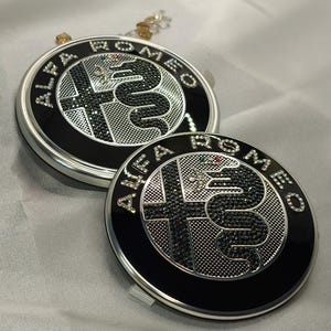 May include: Two black and silver Alfa Romeo emblem badges with a snake and cross design, encrusted with rhinestones. The badges are circular with the brand name around the edge. The background is a light, silky fabric.