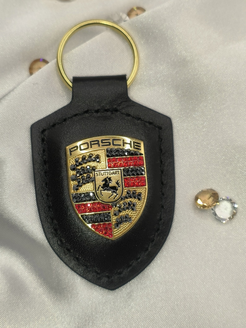 May include: Black leather keychain with a gold Porsche logo featuring red and black rhinestones. The logo says "PORSCHE" and "STUTTGART".