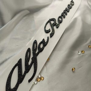 May include: A black, crystal-covered "Alfa Romeo" emblem on a white backing. The cursive text is made of small, dark crystals. The emblem is on a white surface with scattered clear and gold-coloured crystals.