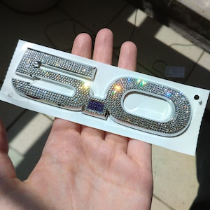 May include: A silver and white car emblem with the number "5.0" in rhinestones. The emblem is on a white backing.