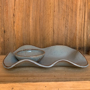 May include: A light blue ceramic serving dish with a wavy design, featuring a small, round bowl. The dish has a speckled texture and a brown rim. The background is a wooden surface.