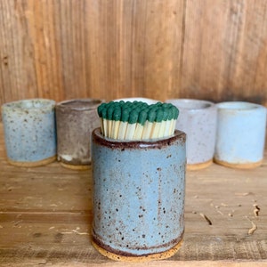 May include: A blue ceramic match striker with a brown rim and a speckled glaze. The striker is filled with matches with green tips.