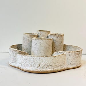 May include: A ceramic centerpiece featuring a low, wide, scalloped-edge bowl with an embossed floral pattern, containing four upright cylindrical holders. The entire piece is finished in a speckled off-white glaze with a visible clay base.