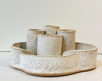 May include: A ceramic centerpiece featuring a low, wide, scalloped-edge bowl with an embossed floral pattern, containing four upright cylindrical holders. The entire piece is finished in a speckled off-white glaze with a visible clay base.