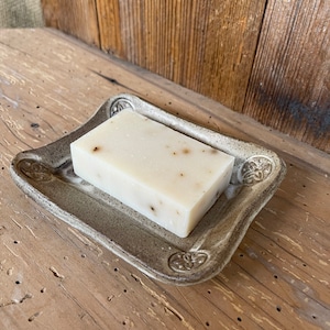 May include: A white bar of soap with flecks of herbs sits on a gray ceramic soap dish with decorative details.