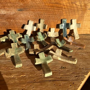 May include: A collection of small, handmade ceramic crosses in various earth-tone colors, including green, beige, and brown. The crosses are arranged on a rustic wooden surface, showcasing their unique textures and shapes. The crosses are a symbol of faith and spirituality.