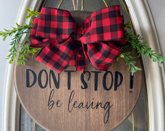 May include: A round wooden door sign with the text "DON'T STOP! be leaving" in black script. It features a red and black buffalo plaid bow and green sprigs. The sign is hanging on a white door.