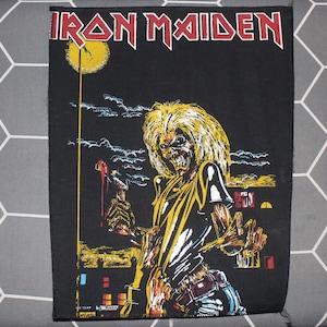 May include: A black Iron Maiden patch with the band's name in red and white lettering. The patch features an illustration of Eddie, the band's mascot, holding a weapon. The artwork includes yellow, red, and white accents against a night sky. Rectangular shape.