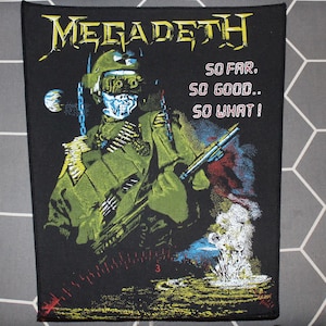 Peut inclure: Écusson noir avec le nom du groupe "Megadeth" en jaune et le texte "SO FAR, SO GOOD.. SO WHAT!". Il présente un soldat squelettique en vert avec une arme, une chaîne et des munitions. Une explosion est représentée en bas à droite.