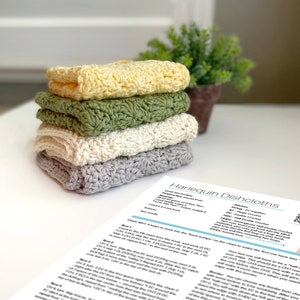 May include: A stack of four crocheted dishcloths in different colors: yellow, green, white, and gray. The dishcloths are made with a textured stitch and have a square shape. The dishcloths are stacked on a white surface with a printed pattern for crocheting dishcloths.
