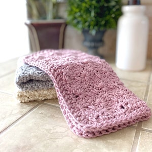 May include: Three crocheted dishcloths in pink, gray, and cream. The dishcloths are folded and stacked on a tiled countertop.