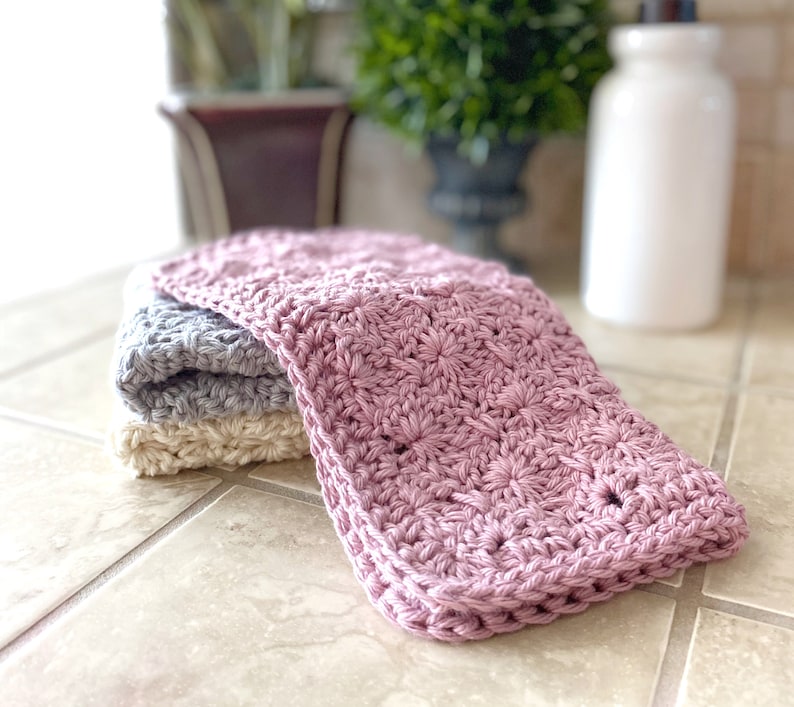 May include: Three crocheted dishcloths in pink, gray, and cream. The dishcloths are folded and stacked on a tiled countertop.