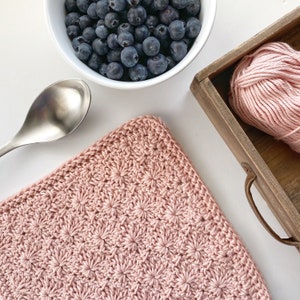May include: A pink crocheted dishcloth with a starburst pattern. The dishcloth is lying on a white surface next to a bowl of blueberries, a spoon, and a wooden box with pink yarn.