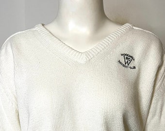 Könnte beinhalten: Weißer V-Ausschnitt-Pullover mit langen Ärmeln und einem kleinen gestickten Logo mit der Aufschrift "Woodstock Racquet Club" mit einem stilisierten "W" in der Mitte.