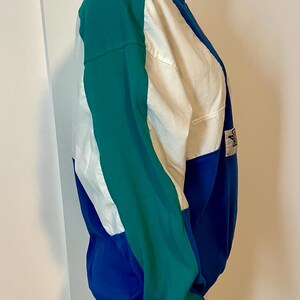 May include: A color-blocked sweatshirt in shades of teal, white, and blue. The long-sleeved top features a crew neck and a small logo patch. The design is a mix of solid colors and geometric shapes, creating a sporty look.