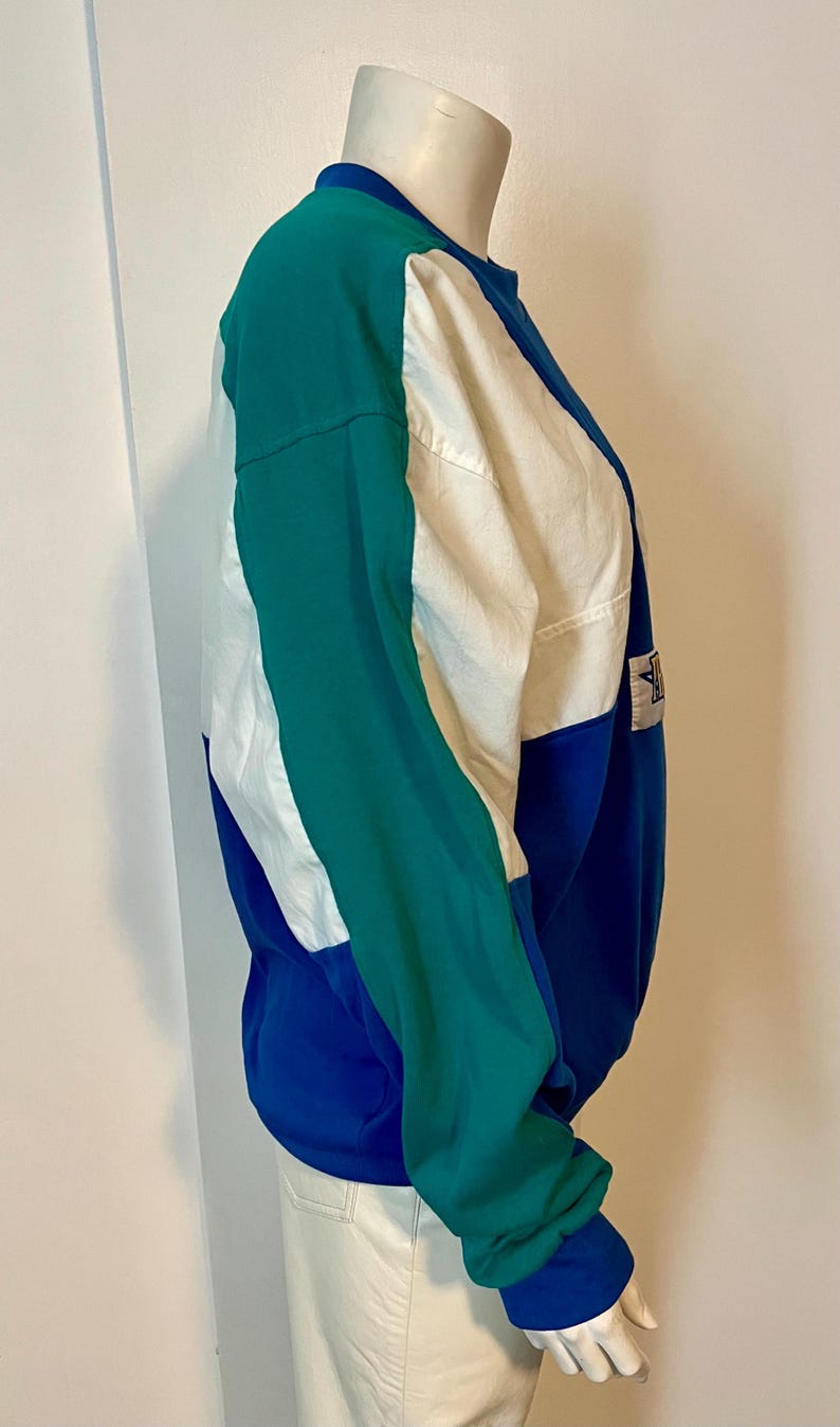 May include: A color-blocked sweatshirt in shades of teal, white, and blue. The long-sleeved top features a crew neck and a small logo patch. The design is a mix of solid colors and geometric shapes, creating a sporty look.