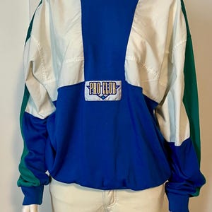 May include: A color-blocked pullover with a blue, white, and green design. The top features a blue center panel with the text "PRO CLUB" on a rectangular patch. The sleeves and lower portion are blue, with green accents on the shoulders.