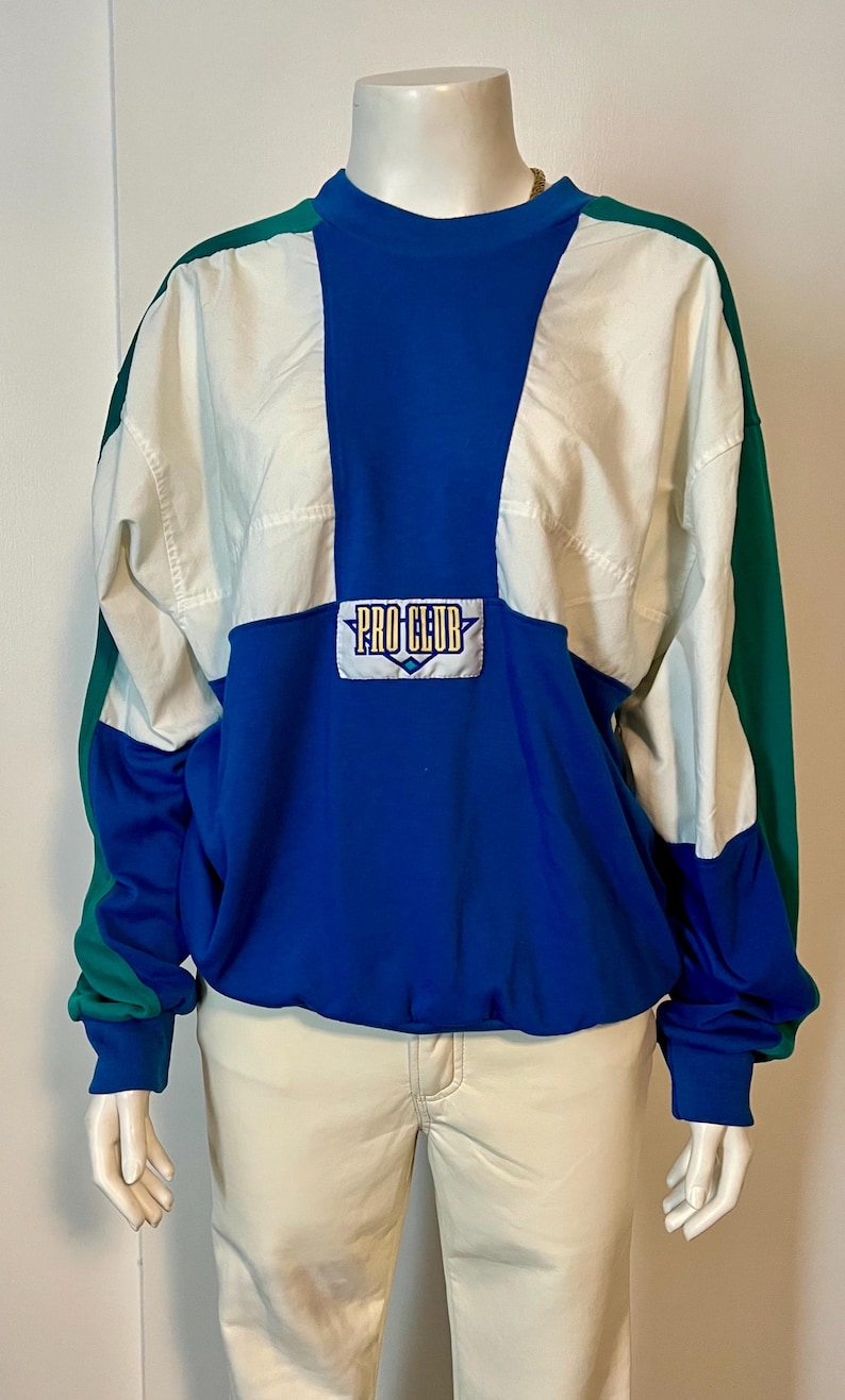 May include: A color-blocked pullover with a blue, white, and green design. The top features a blue center panel with the text "PRO CLUB" on a rectangular patch. The sleeves and lower portion are blue, with green accents on the shoulders.