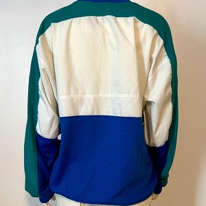 May include: A color-blocked jacket in shades of blue, white, and green. The jacket features a blue collar and cuffs, with a white panel across the chest and a green panel on the shoulders and sleeves. The jacket is displayed on a mannequin.