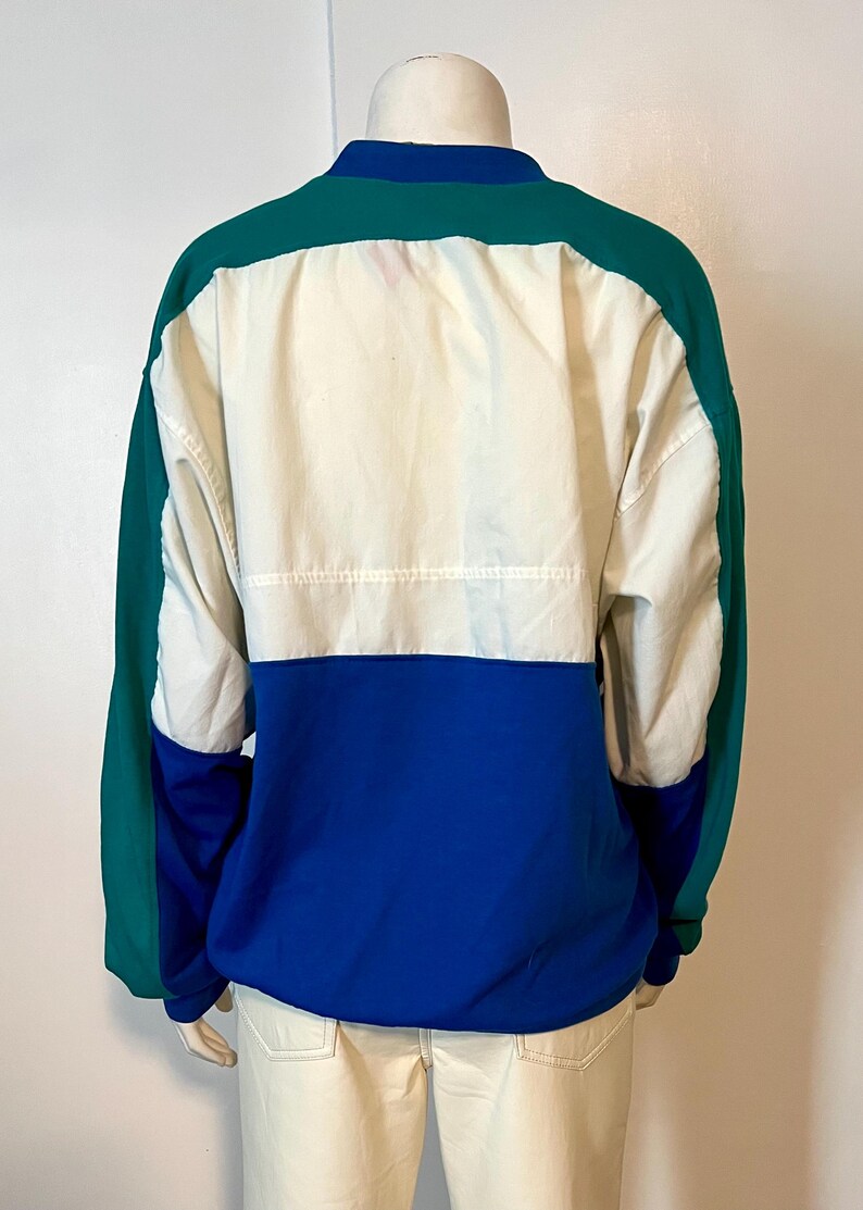 May include: A color-blocked jacket in shades of blue, white, and green. The jacket features a blue collar and cuffs, with a white panel across the chest and a green panel on the shoulders and sleeves. The jacket is displayed on a mannequin.
