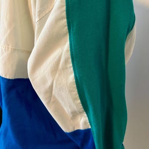 May include: A color-blocked jacket in cream, blue, and green. The jacket features a cream-colored upper section, a blue lower section, and a green sleeve. The design includes a ribbed green sleeve and a contrasting color block design.