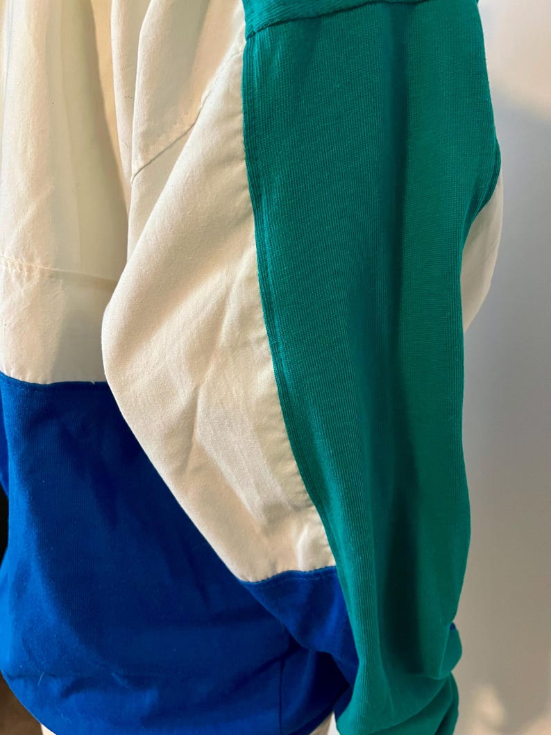 May include: A color-blocked jacket in cream, blue, and green. The jacket features a cream-colored upper section, a blue lower section, and a green sleeve. The design includes a ribbed green sleeve and a contrasting color block design.