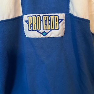 May include: Close-up of a blue and white athletic top. The top features a "PRO CLUB" patch in yellow and blue. The top has a ribbed texture and a classic athletic design.